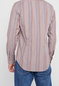 Mand iført en langærmet skjorte med multicolore vertikale striber og blå jeans, set fra ryggen mod en ensfarvet baggrund.