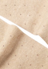 Bēša adīta šalle ar mīkstu struktūru, rotāta ar izkaisītiem maziem metāliskiem kniedēm akcentam. Dizainā ir vienkāršs, elegants raksts.