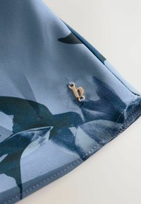 Tessuto floreale blu con una texture satinata, caratterizzato da un accento in metallo dorato a forma della lettera "b" vicino all'orlo.
