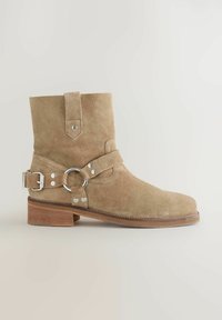 Beiger Wildleder-Stiefelette mit abgerundeter Spitze und flachem Holzabsatz. Verfügt über einen Riemen mit einem runden Metallring und Schnallen zur Anpassung.