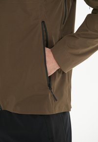 Veste légère marron avec poches zippées noires et texture lisse. Présente une coupe ajustée et des manches taillées.