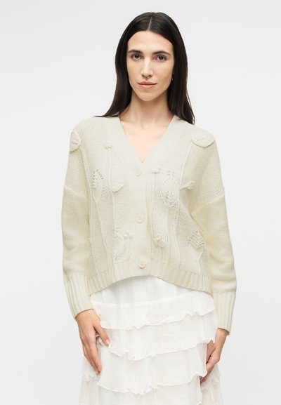 Γυναίκα με μακριά σκούρα μαλλιά, ντυμένη με εκρού πλεκτή ζακέτα με υφασμένα σχέδια φύλλων πάνω από λευκή στιβάδα φούστας, που στέκεται μπροστά σε απλό φόντο.