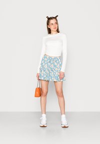 Saia azul estampada com margaridas brancas, com bainha em camadas, combinada com uma blusa branca de mangas longas e ténis brancos, segurando uma bolsa laranja.