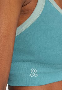 Sujetador deportivo de color azul verdoso con un ribete en un verde claro que contrasta. Presenta un logo blanco en la parte inferior y una textura suave.