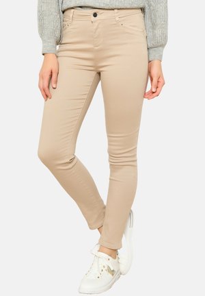 Kvinde iført beige slim fit bukser, grå strikket sweater og hvide sneakers, står med det ene ben krydset over det andet.