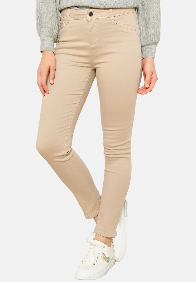 Femme portant un pantalon beige slim, un pull en maille gris et des baskets blanches, debout avec une jambe croisée sur l'autre.