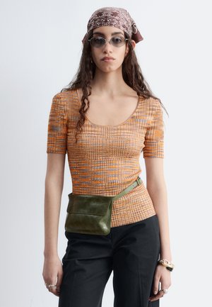 Jeune femme portant un foulard à motifs, des lunettes de soleil ovales, un top orange à rayures à manches courtes, un pantalon noir et une sacoche verte à la taille.