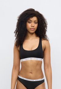 Σετ μαγιό μπικίνι σε μαύρο χρώμα με σουτιέν bralette με στρογγυλή λαιμόκοψη και αντίστοιχο κάτω μέρος. Διαθέτει λευκή ταινία με τη φράση "TOMMY HILFIGER". Το ύφασμα φαίνεται λείο και ελαστικό.