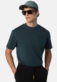 Uomo che indossa una maglietta North Sails di colore verde scuro, occhiali da sole neri, cappellino grigio e pantaloni neri con le mani nelle tasche, su uno sfondo neutro.