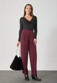 Kobieta ubrana w czarną koronkową bluzkę z dekoltem w kształcie V, wysokie bordowe spodnie, czarne botki za kostkę, trzymająca dużą czarną torebkę z złotymi łańcuszkami.