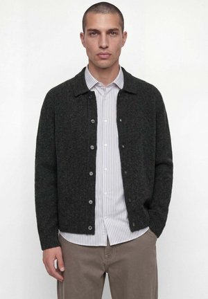 Homme portant un cardigan sombre boutonné sur une chemise rayée blanche et grise et un pantalon beige, debout devant un fond blanc uni.