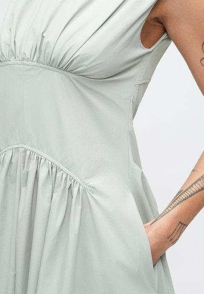 Svetlozelené šaty bez rukávov s nazberanými detailmi okolo pása a splývavou sukňou. Materiál pôsobí hladko a ľahko.
