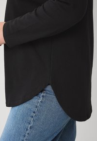 Haut noir à manches longues avec un ourlet arrondi, présentant une texture douce et un design superposé, associé à un jean en denim bleu.