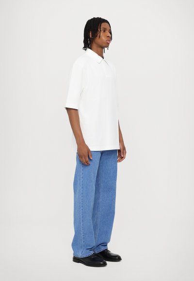 White polo shirt with a collar and short sleeves, paired with light blue wide-leg jeans and black shoes, shown from the side.