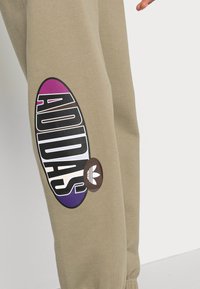 Beige sporta bikses ar lielu melnu ovālu logotipu, uz kura ir "ADIDAS", un purpura krāsas gradientu. Iekļauta brūna akcenta detaļa ar Adidas trefoil.