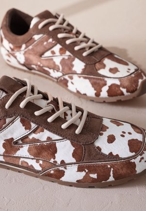 Sneakers mit braun-weißem Kuhfleckenmuster aus Wildleder und Kunstfell, mit hellbraunen Schnürsenkeln und Gummisohle. Abgerundete Zehenspitzengestaltung.