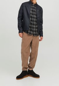 Veste noire en simili cuir avec col montant, chemise à carreaux en-dessous, pantalon en velours côtelé beige, et bottines en daim noir avec semelle beige.