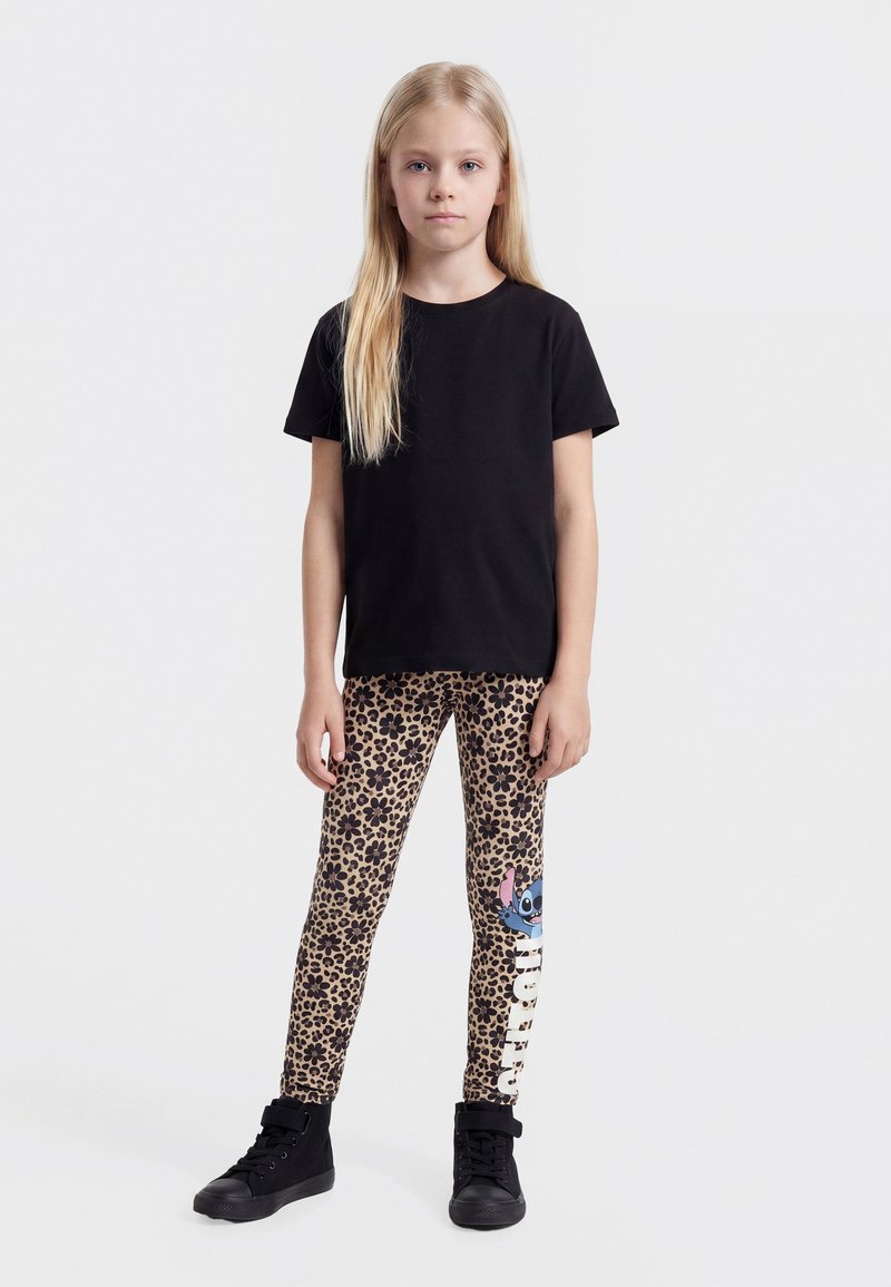 Fille debout face à l’avant, portant un t-shirt noir, des leggings imprimés léopard avec un personnage de dessin animé bleu et des baskets montantes noires.