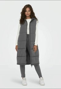 Colete puffer cinza com capuz, fecho de correr à frente e padrão retangular acolchoado, usado sobre uma camisa branca e calças cinzas, combinado com ténis brancos.