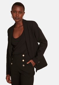 Blazer nero con taglio sartoriale, abbinato a una canotta nera e pantaloni a vita alta con dettagli di bottoni dorati. Tessuto liscio e design pulito.