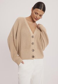 Beżowy sweter z dzianiny z głębokim dekoltem w kształcie litery V, fakturowanym wzorem i dużymi brązowymi guzikami, w połączeniu z białymi spodniami. Luźny krój.