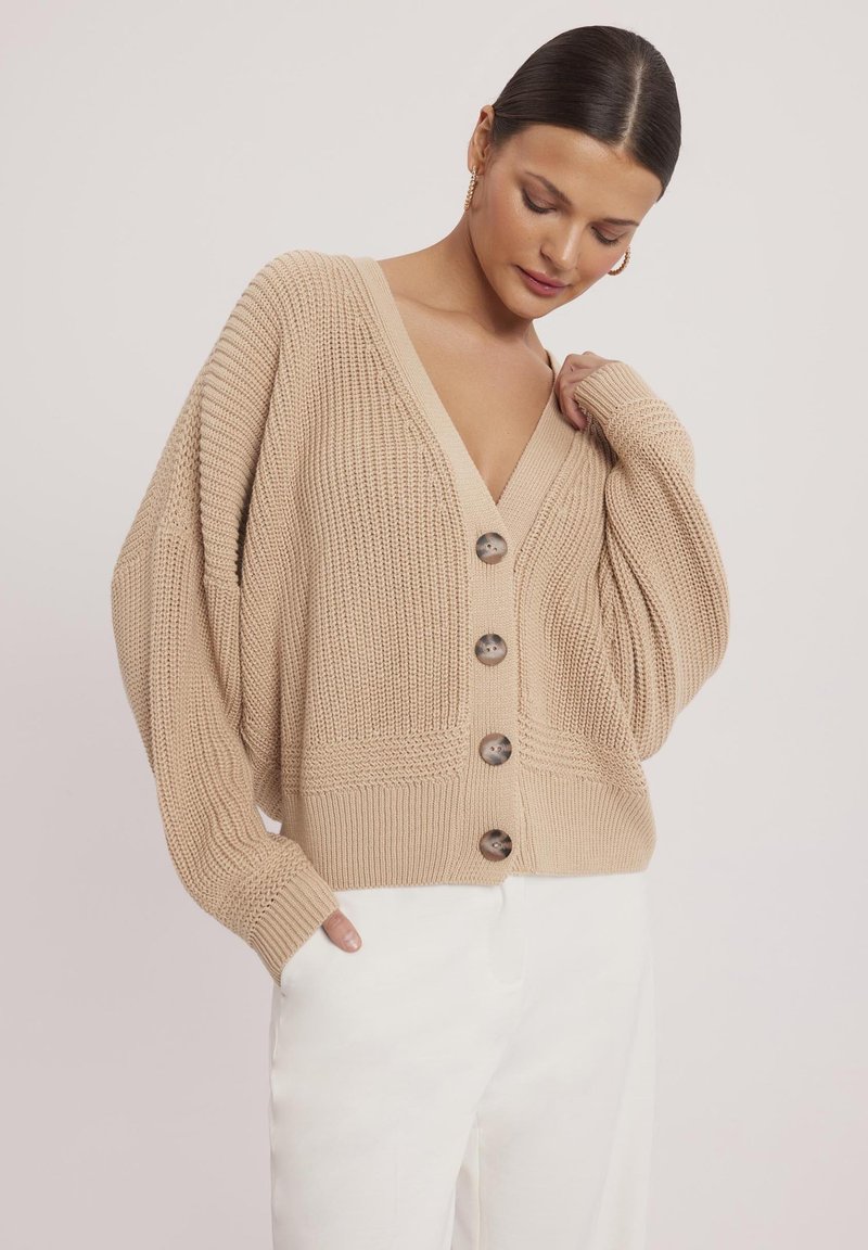 Beżowy sweter z dzianiny z głębokim dekoltem w kształcie litery V, fakturowanym wzorem i dużymi brązowymi guzikami, w połączeniu z białymi spodniami. Luźny krój.