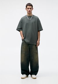 T-shirt in cotone oversize verde scuro abbinato a jeans a gamba larga con strappi e sneaker bianche. Collana a catena in argento visibile.