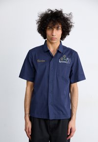 Marineblauw kortemouwenshirt van katoen met een linkerklepzak, voorzien van groene borduring van bloemen en tekst aan de voorkant.