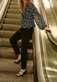 Une femme portant un chemisier fleuri à manches longues et un pantalon noir se tient sur un escalator descendant, tenant la rampe d'une main.