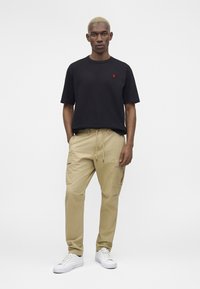 Άνδρας με κοντά ξανθά μαλλιά, φοράει μαύρο κοντομάνικο πουκάμισο, μπεζ παντελόνι cargo και λευκά αθλητικά παπούτσια, στέκεται με τα χέρια εν μέρει στις τσέπες.