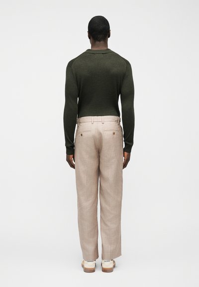 Άνδρας που κοιτάζει προς τα πίσω, φοράει σκούρο πράσινο ριμπ πουλόβερ, μπεζ παντελόνι με ζώνη και λευκά αθλητικά παπούτσια σε απλό φόντο.