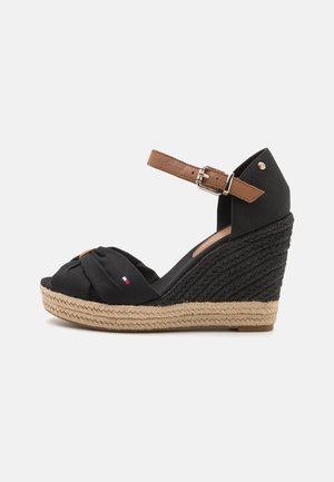 Sandales à talon noir avec une tige en tissu noué, une sangle de cheville beige et des détails en jute tressé sur le talon. Accentué d'un petit logo en forme de drapeau.