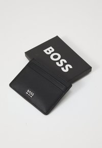 Melnā kartes turētāja izgatavota no teksturēta materiāla ar vairākām kabatām. Ietver melnu kastīti ar izteiksmīgu baltu "BOSS" zīmolu.