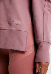 Detailní záběr ruky u boků, oblečené v mauve mikině značky Adidas Stella McCartney a odpovídajících legínách s texturovanými a hladkými tkaninovými panely.