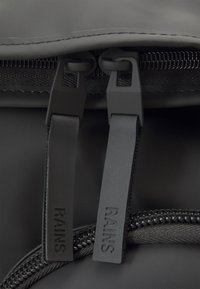 Detail des Reißverschlusses eines schwarzen Rucksacks mit zwei Zugbändern, die jeweils mit "RAINS" geprägt sind. Das Material hat eine glatte, matte Oberfläche und eine robuste Naht.