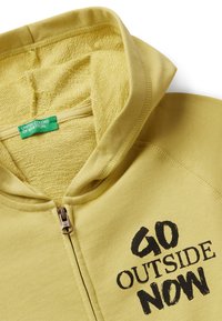 Geel ritsvest met een gestructureerde binnenkant, voorzien van de zwart gedrukte tekst "GA NAAR BUITEN NU" op de voorkant. Etiket zegt "UNITED COLORS OF BENETTON."