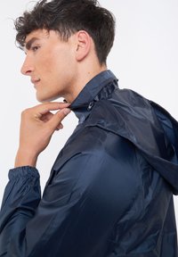 Giacca leggera blu navy con colletto rialzato, dettagli con zip e polsini elastici. Il materiale sembra liscio e resistente all'acqua.