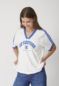 T-shirt bianca con accenti blu, scollo a V e scritta grafica "CO'COUTURE LOVE & JOY." Maniche corte con strisce blu ai lati.
