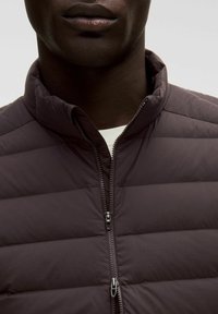 Giacca trapuntata marrone scuro con colletto alto e zip centrale, caratterizzata da una texture liscia e dettagli di cucitura orizzontale.