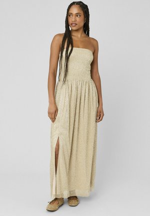 Femme aux longues tresses portant une robe longue beige sans bretelles à texture avec une fente latérale et des chaussures plates assorties ornées.