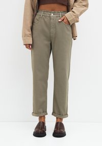 Pantalon ample vert olive avec une ceinture élastique, ourlets retroussés, poches latérales et un détail bouton à l'avant, assorti à des chaussures marron à enfiler.