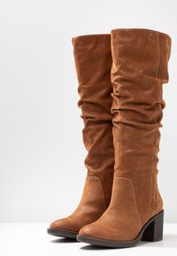 Tan leather knee-high boots with a slouchy design, textured surface, and chunky heel. Features a rounded toe and visible stitching detail.