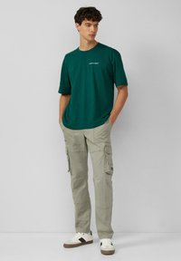 Donkergroene t-shirt met korte mouwen en een ronde hals, gecombineerd met lichtbeige cargo-broeken met meerdere zakken. Casual witte sneakers.
