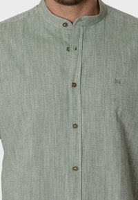 Camisa de lino de color verde claro con cuello tipo banda, con un tejido texturizado, cinco botones de latón y un pequeño logotipo bordado en el pecho.