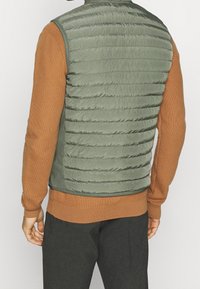 Padded green vest featuring horizontal quilting, with a contrasting brown textured long-sleeve shirt underneath. Dark pants visible.