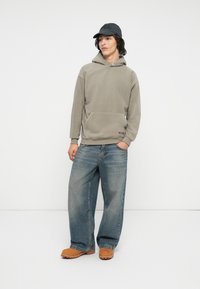 Lys olivengrønn fleecehettegenser med kengurulomme, kombinert med løstsittende blå denimjeans og beige slip-on sko. En svart caps fullfører antrekket.