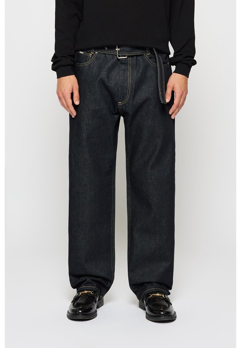 Personne portant un jean en denim foncé ample avec une ceinture noire et des mocassins noirs avec des détails en chaîne dorée.