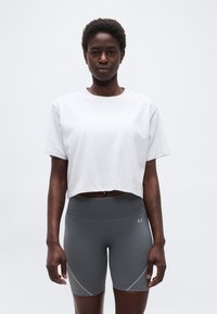 Γυναίκα με κοντά μαλλιά που φοράει λευκό crop top και γκρι αθλητικές σορτς με ψηλή μέση, στέκεται μπροστά σε απλό φόντο.