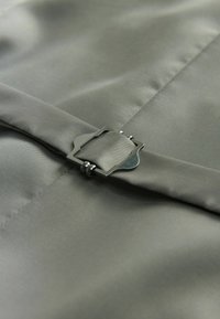 Tissu gris avec une texture lisse, comportant une boucle en métal et un détail de sangle. Le design présente un bord replié et des coutures soignées.