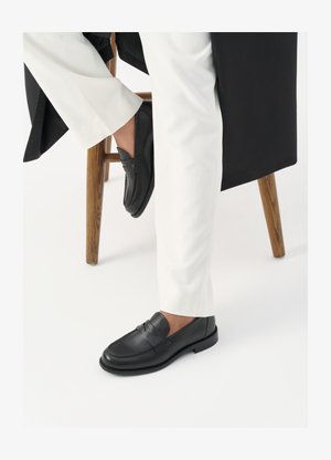 Black leather loafers with a smooth texture and a wide strap detail, worn with white tailored pants. The heel features a flat design.